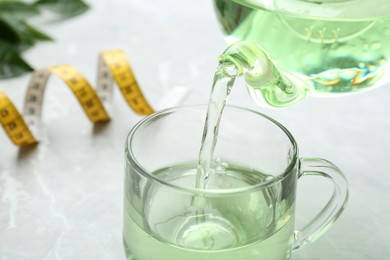 Pouring herbal diet tea into cup at light table with measuring tape, closeup