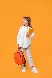Happy schoolgirl with backpack and book on orange background