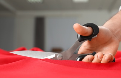 Photo of Professional tailor cutting red fabric with scissors in workshop, closeup