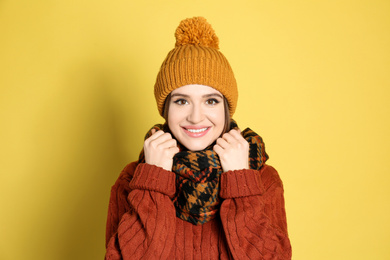 Young woman wearing warm sweater, scarf and hat on yellow background. Winter season