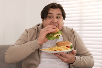 Photo of Overweight man eating tasty burger on sofa at home