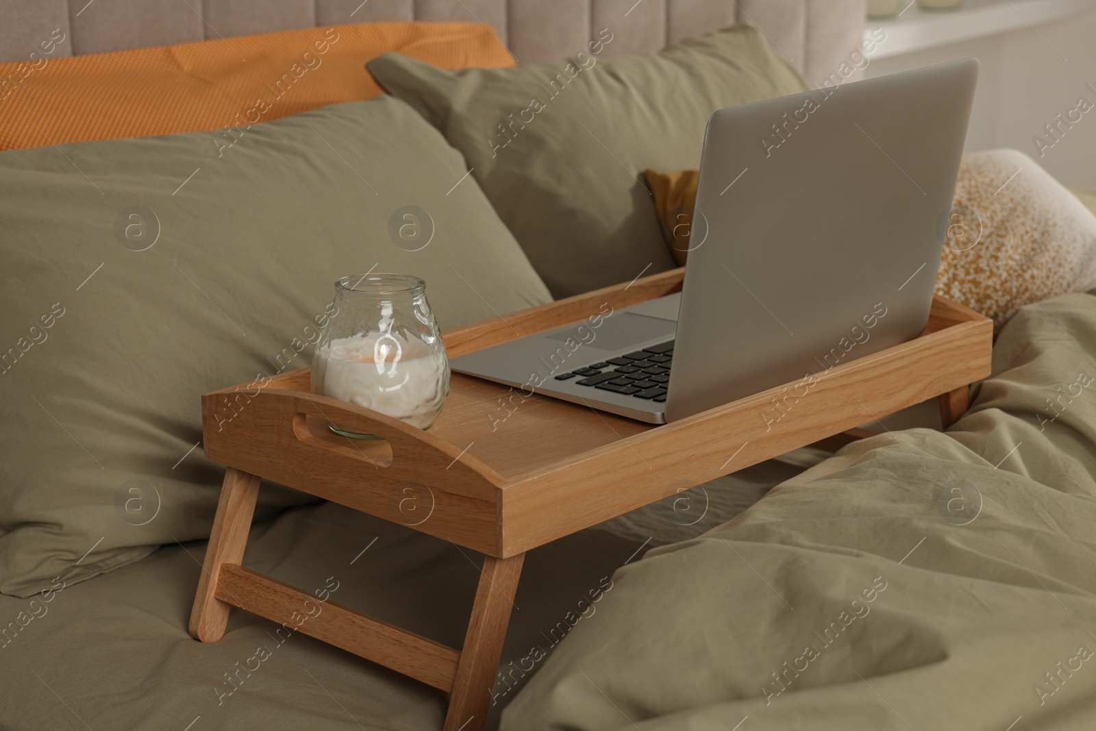 Photo of Wooden tray with modern laptop and burning candle on bed indoors