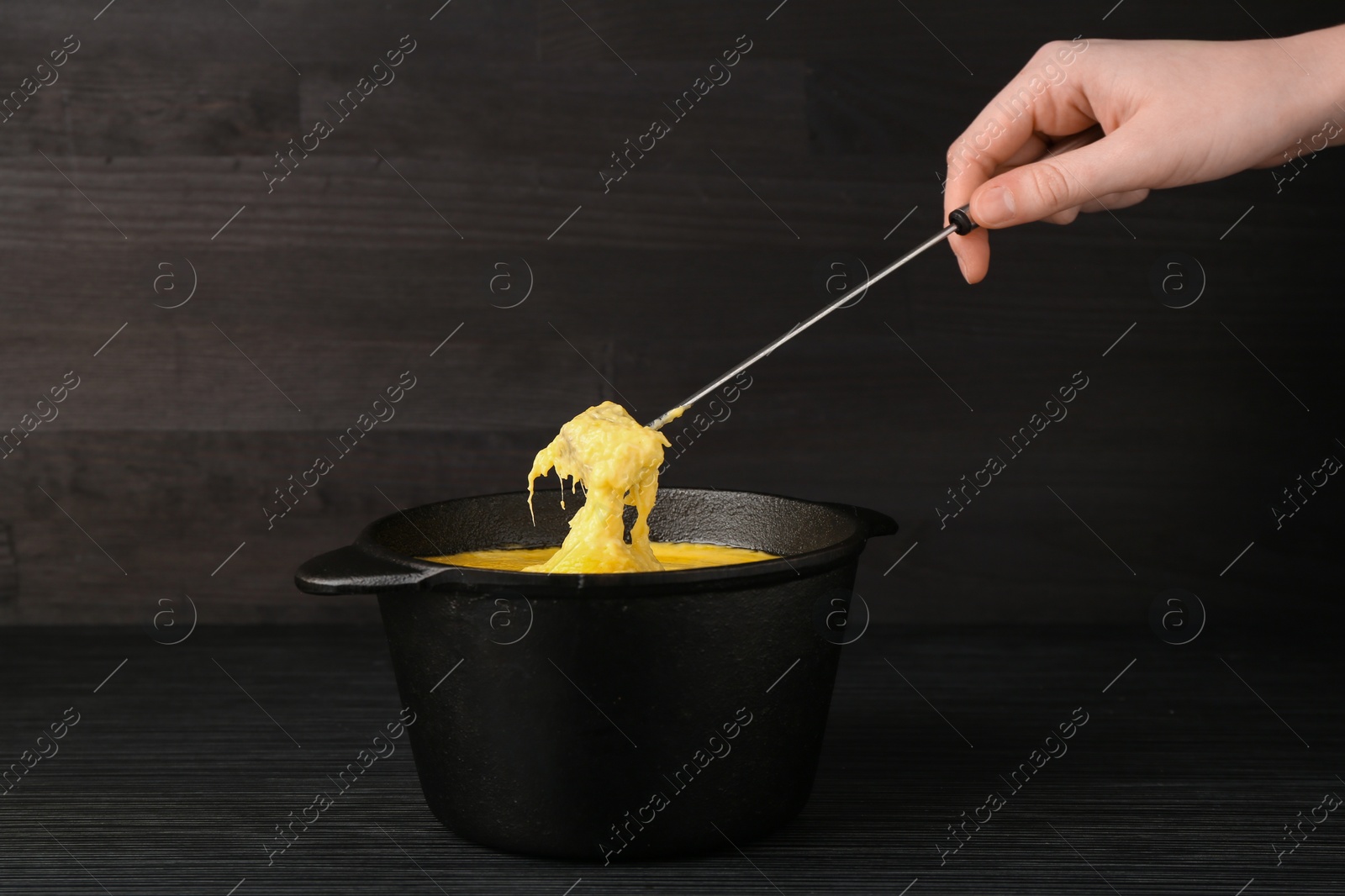 Photo of Woman dipping piece of bread into fondue pot with melted cheese at black wooden table, closeup