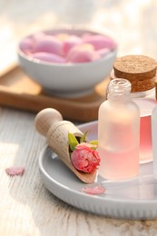 Bottles of rose essential oil and flowers on white wooden table, closeup