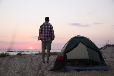 Photo of Man enjoying sunset near camping tent on beach, back view