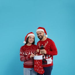Photo of Happy young couple in Christmas sweaters and Santa hats taking gift from stocking on light blue background