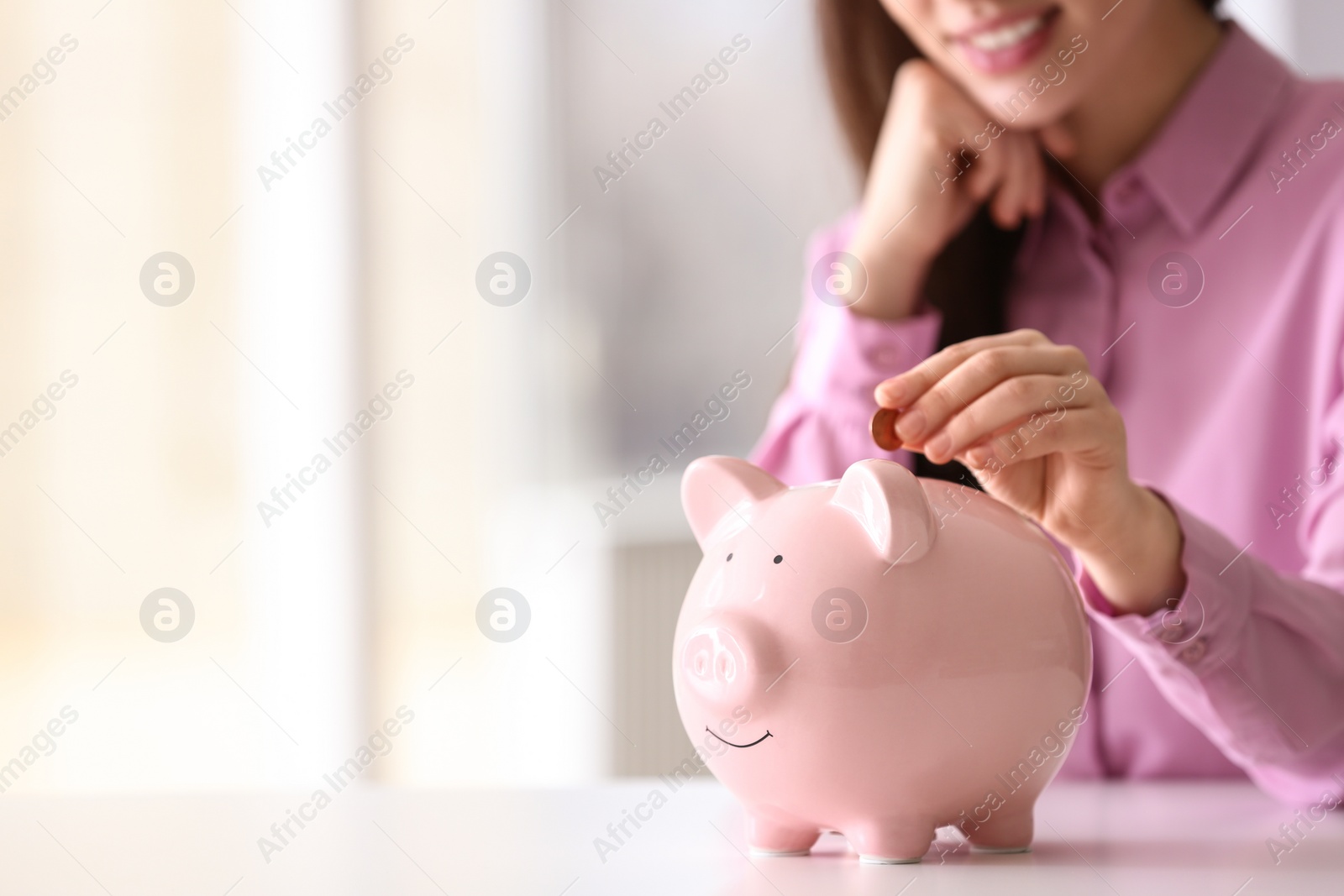 Photo of Woman putting coin into piggy bank at table indoors, closeup. Space for text