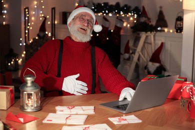 Santa Claus using laptop and laughing at his workplace in room decorated for Christmas