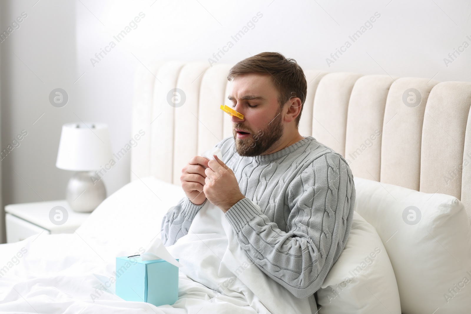 Photo of Young man with clothespin suffering from runny nose in bed indoors