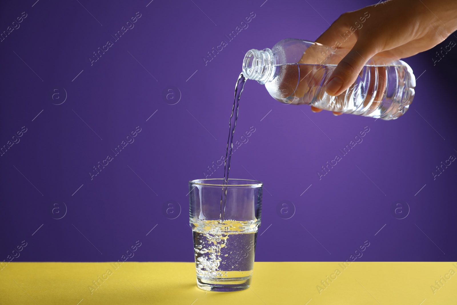 Photo of Woman pouring water from bottle into glass on yellow table against purple background, closeup