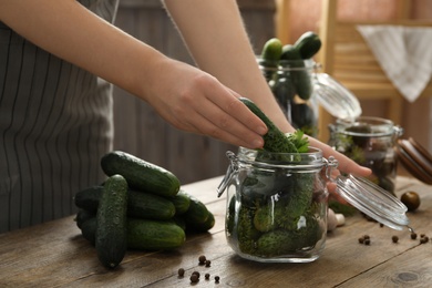 Photo of Woman putting cucumber into glass jar at wooden kitchen table, closeup. Pickling vegetables