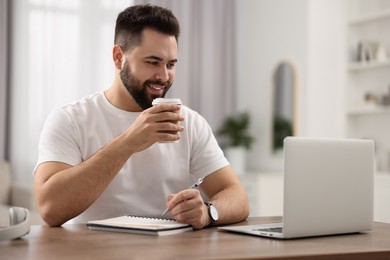 Photo of Young man with cup of coffee watching webinar at table in room