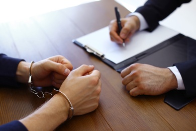 Photo of Police officer interrogating criminal in handcuffs at desk indoors