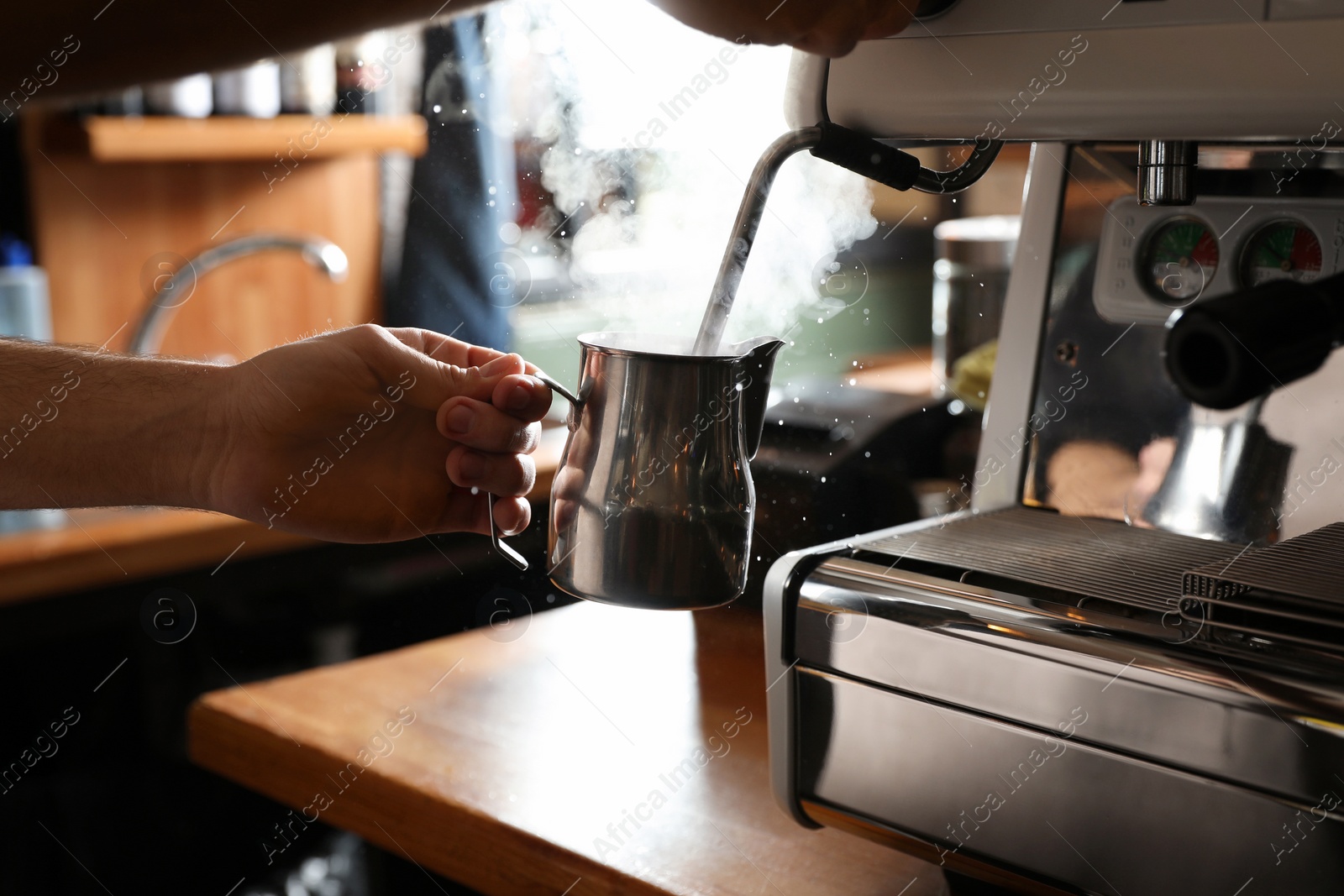 Photo of Barista steaming milk in metal jug with coffee machine wand at bar counter, closeup