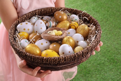 Photo of Little girl holding wicker basket full of Easter eggs outdoors, closeup