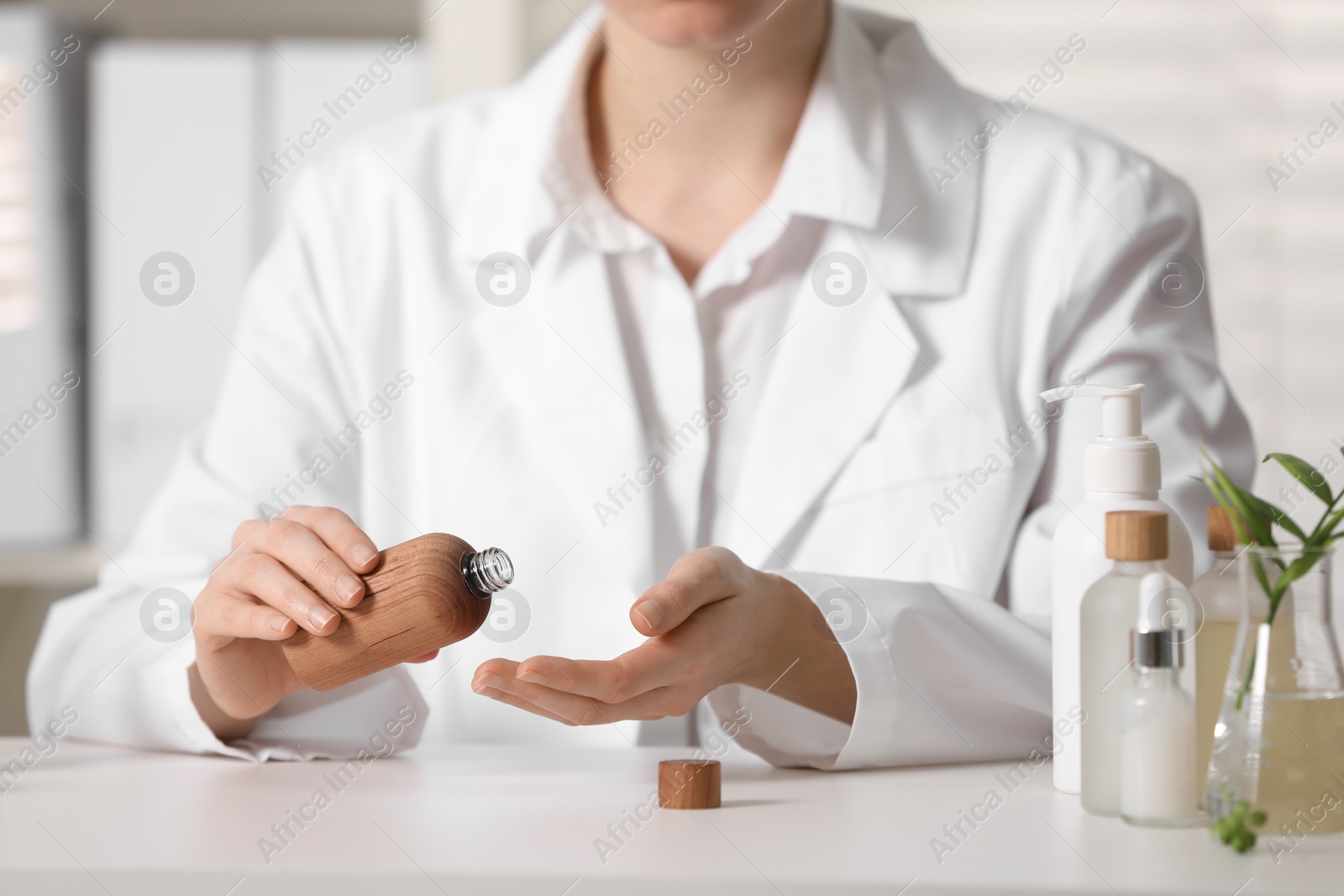 Photo of Dermatologist with bottle testing cosmetic product at white table indoors, selective focus