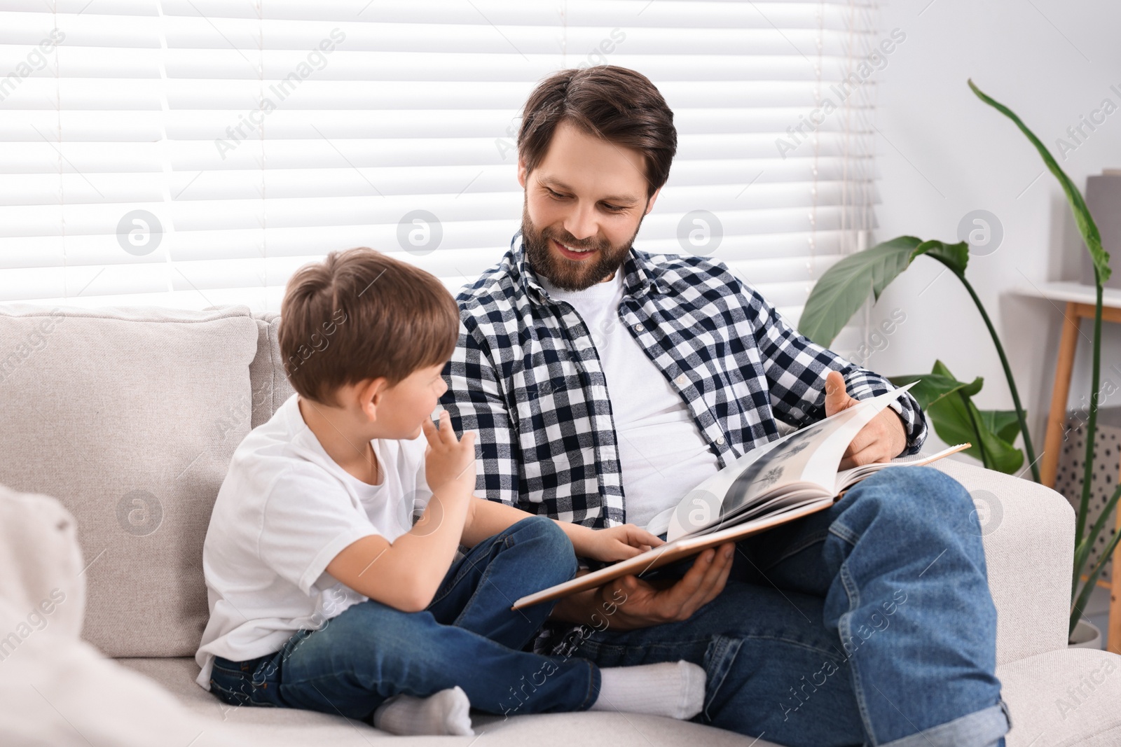 Photo of Happy dad and son reading book together on sofa at home