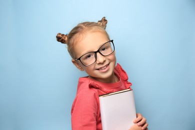 Cute little girl with glasses and textbook on light blue background