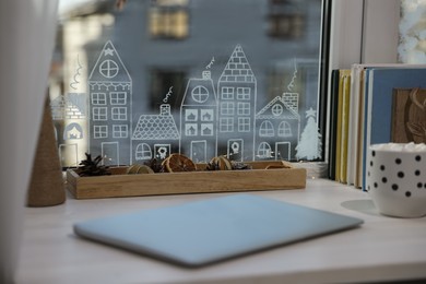 Photo of Laptop and hot drink on white wooden table near window with beautiful drawing at home. Christmas decor