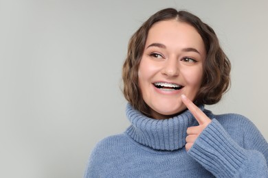 Happy woman in warm sweater pointing at braces on her teeth against grey background. Space for text