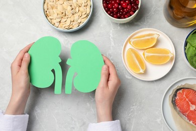 Photo of Woman holding paper cutout of kidneys near different products on light grey table, top view