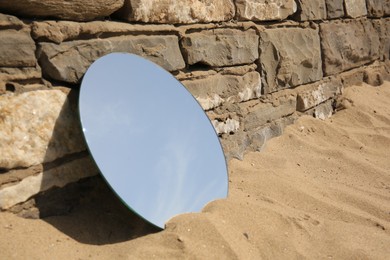 Round mirror reflecting sky on sand near stone wall outdoors, space for text