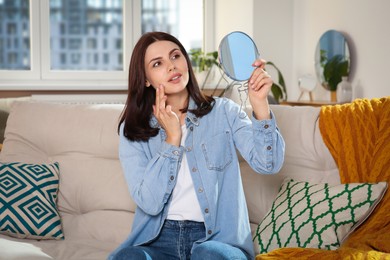 Photo of Young woman looking in mirror and squeezing pimple on sofa at home. Hormonal disorders