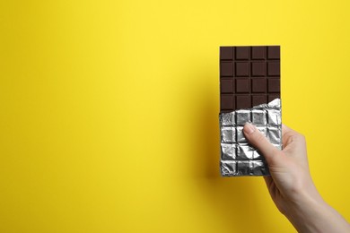 Woman holding delicious dark chocolate bar on yellow background, closeup. Space for text