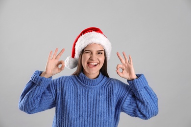 Photo of Pretty woman in Santa hat and blue sweater showing OK gesture on grey background
