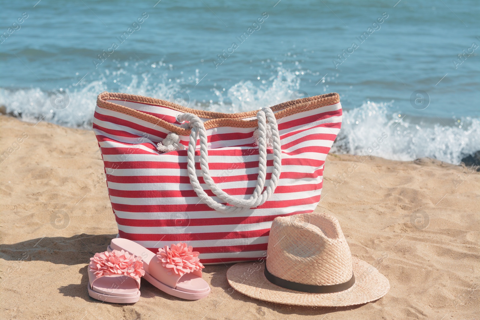 Photo of Stylish striped bag with straw hat and slippers on sandy beach near sea