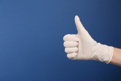 Person in medical gloves showing thumb up on blue background, closeup of hand. Space for text