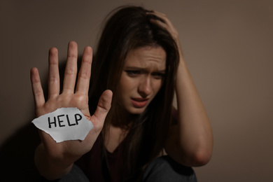 Abused young woman with sign HELP near beige wall, focus on hand. Domestic violence concept