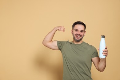 Happy man holding thermo bottle and showing arm on beige background, space for text