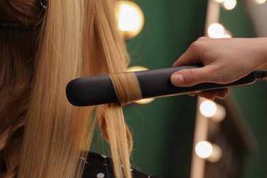 Photo of Stylist curling woman's hair with flat iron in salon, closeup