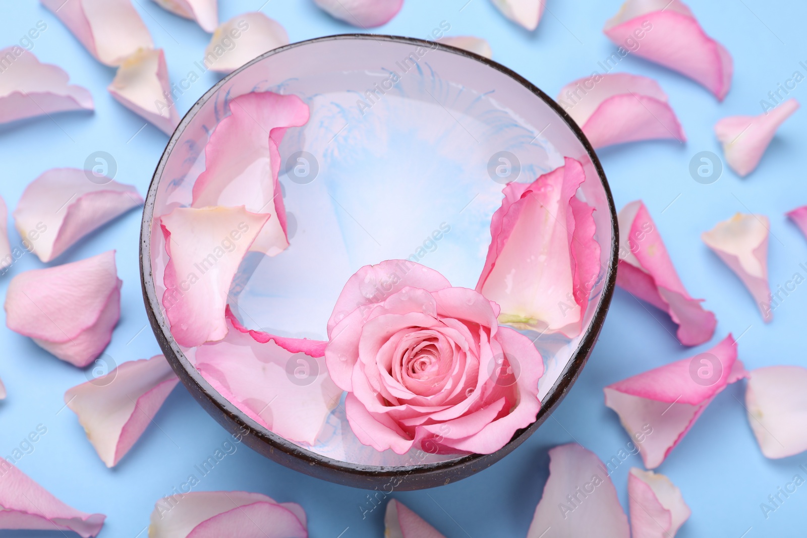 Photo of Bowl with water and rose petals on light blue background, above view. Spa composition