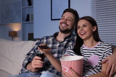 Photo of Happy couple watching show at home in evening. Woman holding popcorn and changing TV channels with remote control