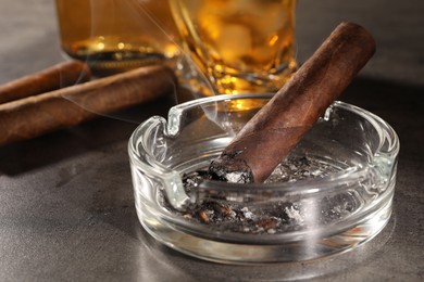 Photo of Smoldering cigar, ashtray and whiskey on grey table, closeup