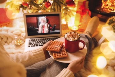 MYKOLAIV, UKRAINE - DECEMBER 23, 2020: Woman with cocoa and wafers watching The Christmas Chronicles movie on laptop near fireplace at home, closeup. Cozy winter holidays atmosphere