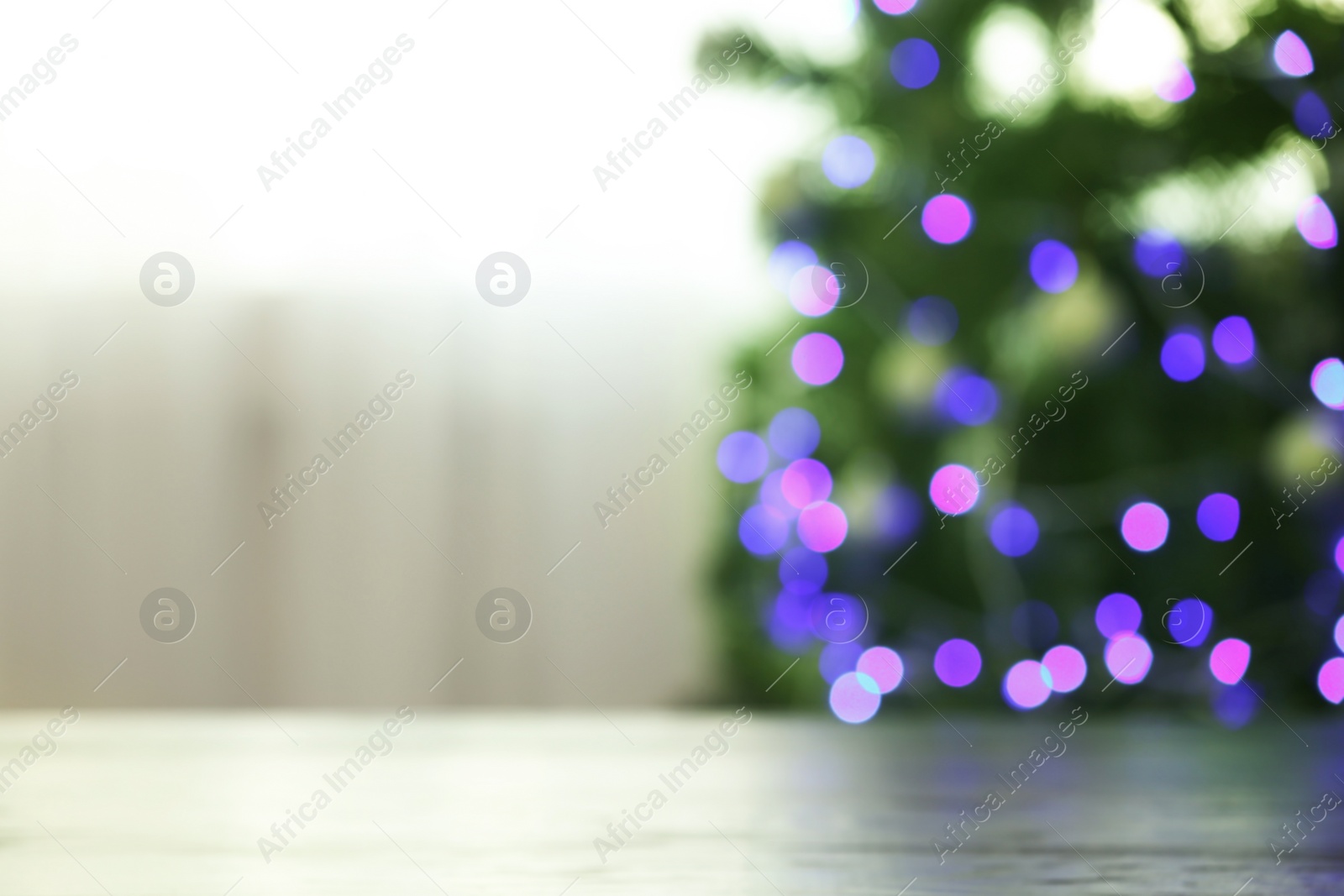 Photo of Blurred view of fir tree with glowing Christmas lights near table indoors