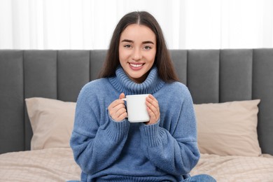 Photo of Happy young woman holding white ceramic mug on bed at home