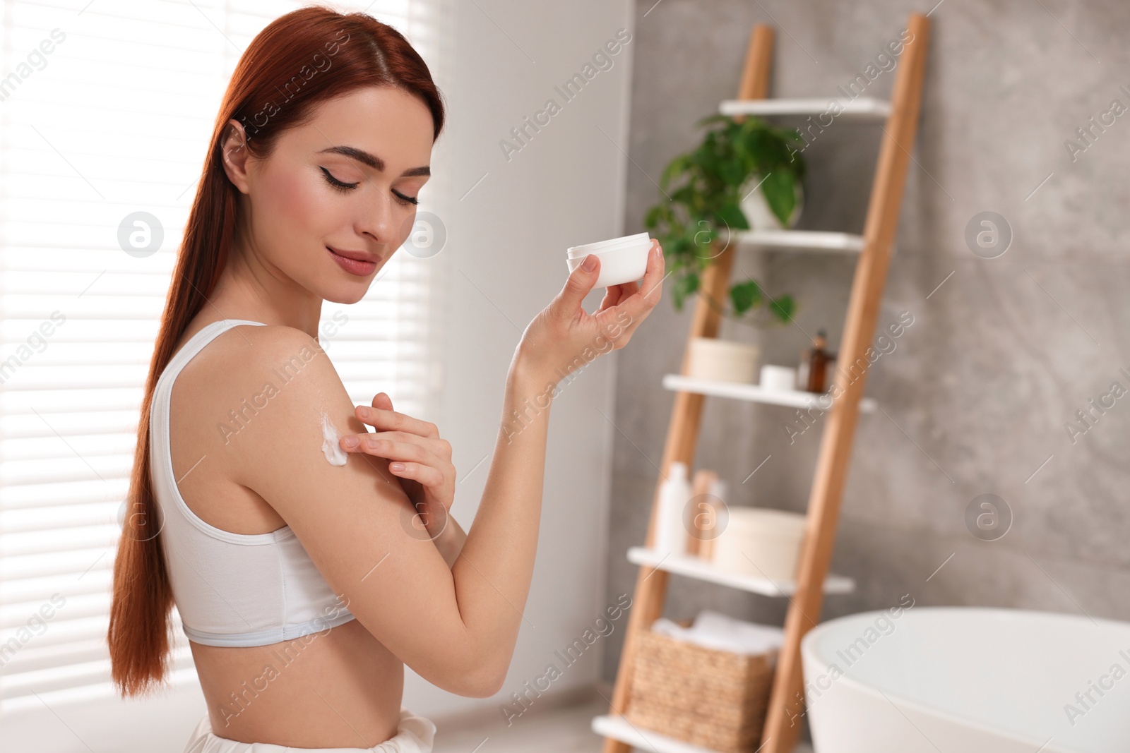 Photo of Beautiful young woman applying body cream onto shoulder in bathroom, space for text