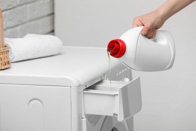 Woman pouring fabric softener from bottle into washing machine near light grey wall, closeup