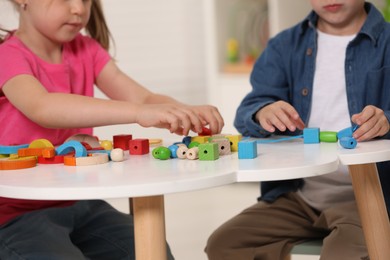 Little children playing with wooden pieces and string for threading activity at white table indoors, closeup. Developmental toys