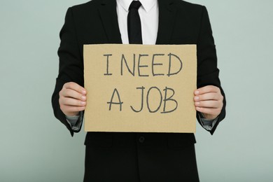 Unemployed man holding sign with phrase I Need A Job on light grey background, closeup