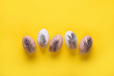 Many painted Easter eggs with feathers on yellow background, flat lay