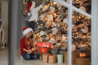 Photo of Cute little children near Christmas tree in living room, view through window