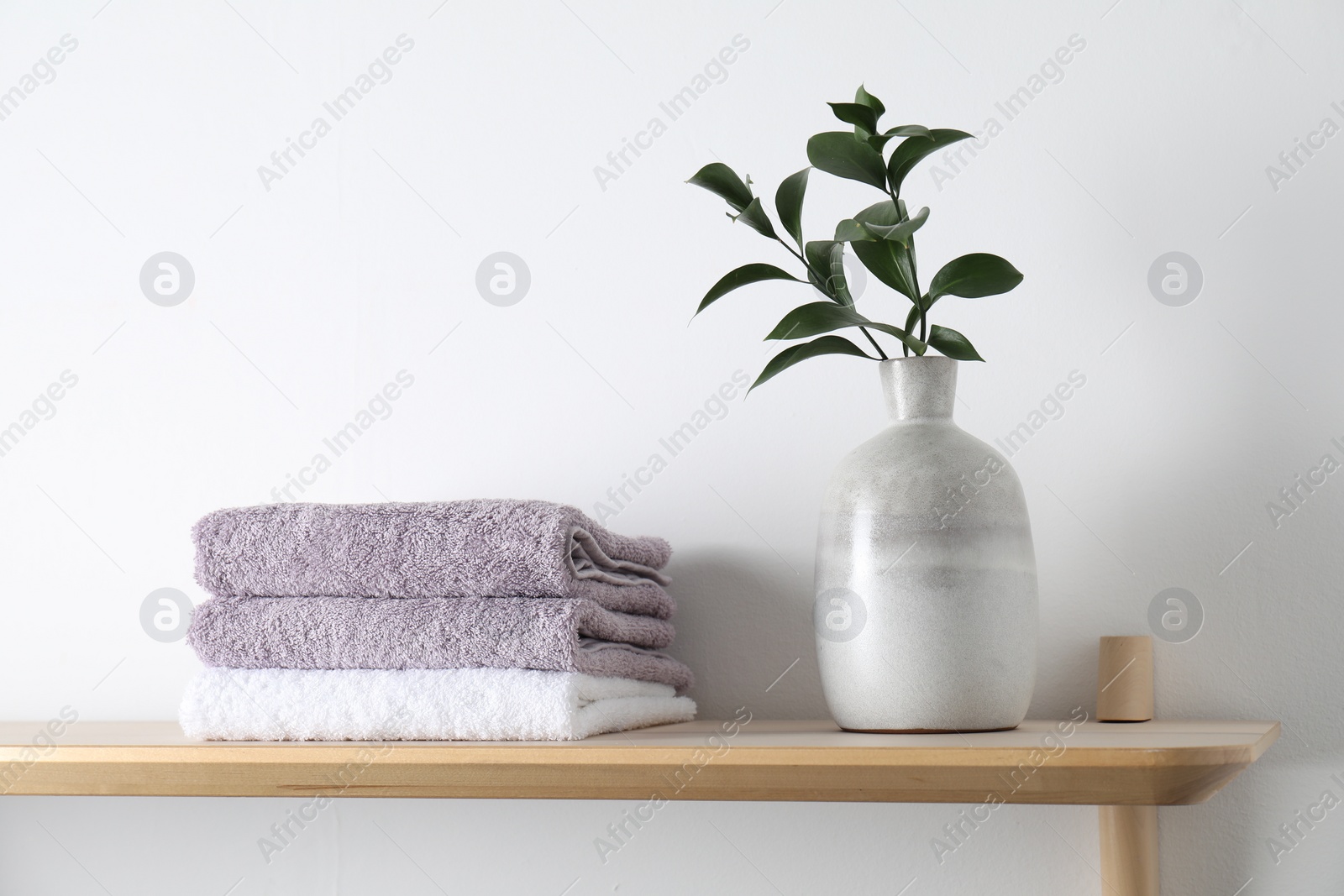 Photo of Stacked terry towels and green branches in vase on wooden shelf near white wall