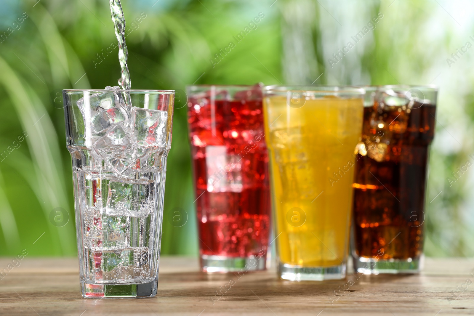 Photo of Pouring refreshing soda water into glass with ice cubes on wooden table outdoors