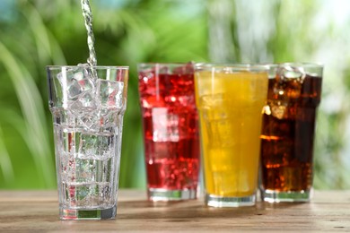 Pouring refreshing soda water into glass with ice cubes on wooden table outdoors