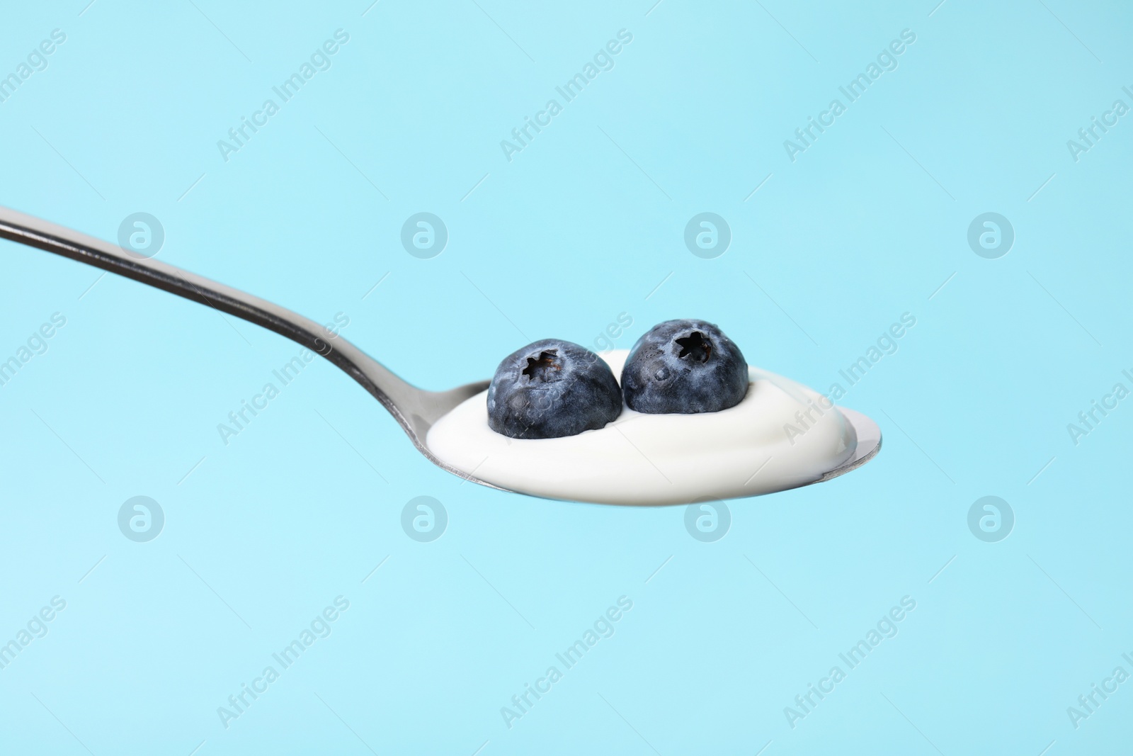 Photo of Spoon with yogurt and blueberries on light blue background, closeup